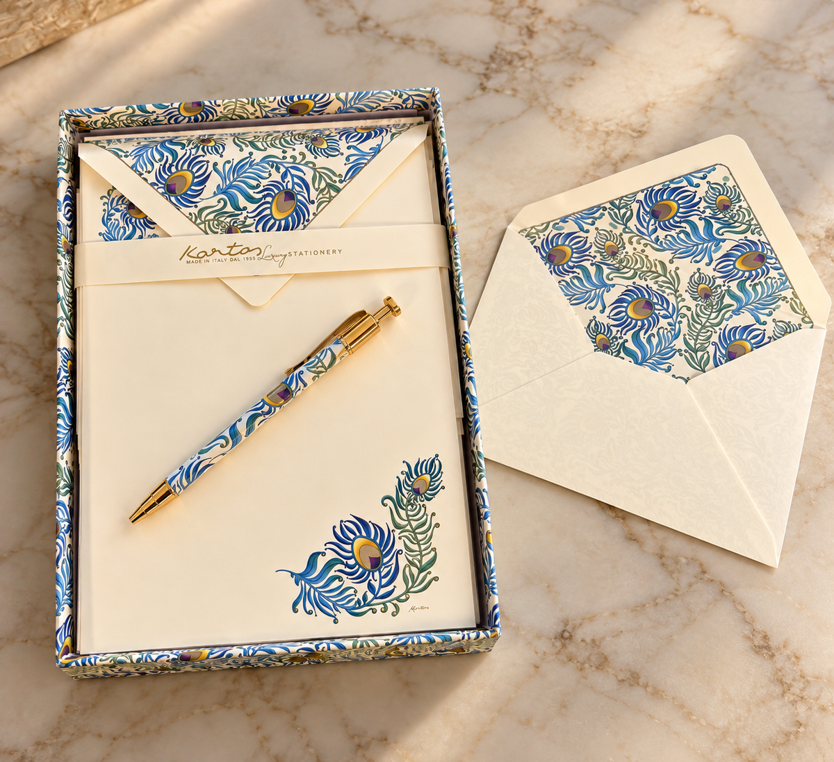 A boxed stationery set on a marble surface, featuring a matching pen, card, and envelope with a blue and yellow floral pattern and a peacock illustration. The open box shows the interior lined with the same patterned paper, and an additional matching envelope lies beside it.
