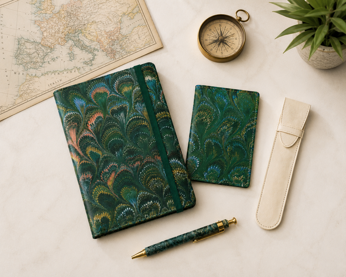 A vintage-inspired travel journal with a rich teal and green peacock-feather pattern, paired with a matching smaller notebook, a coordinating pen, and a cream pen sleeve. A vintage map, brass compass, and potted greenery create an elegant travel-themed still life.