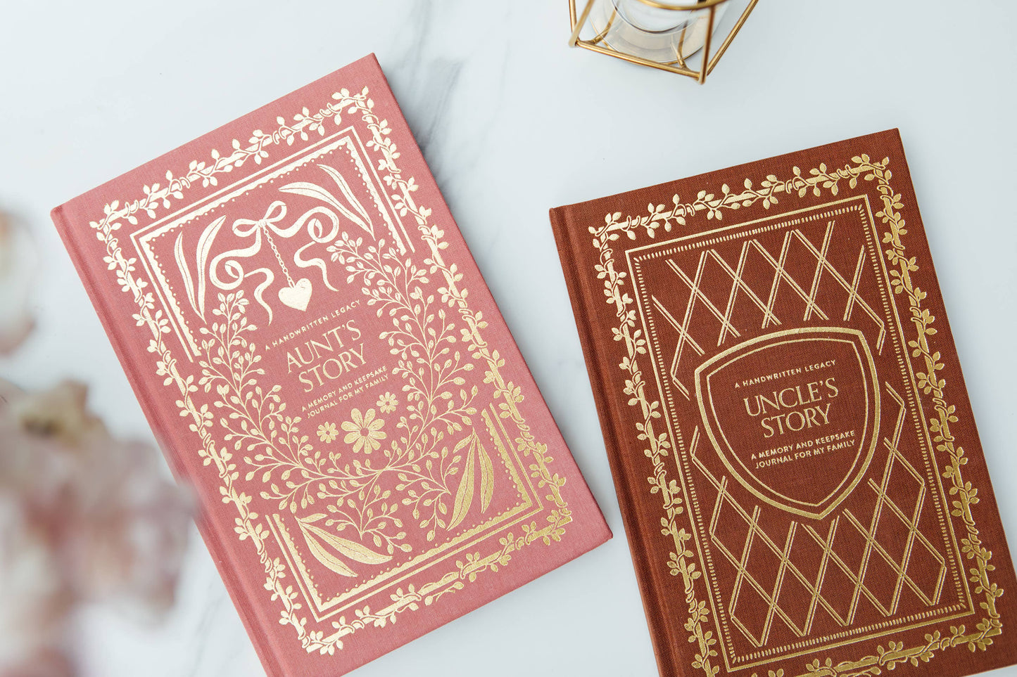 Two decorative hardcover books, 'Aunt's Story' in dusty pink and 'Uncle's Story' in dark brown, both featuring ornate gold foil borders and titles, arranged on a light marble surface.