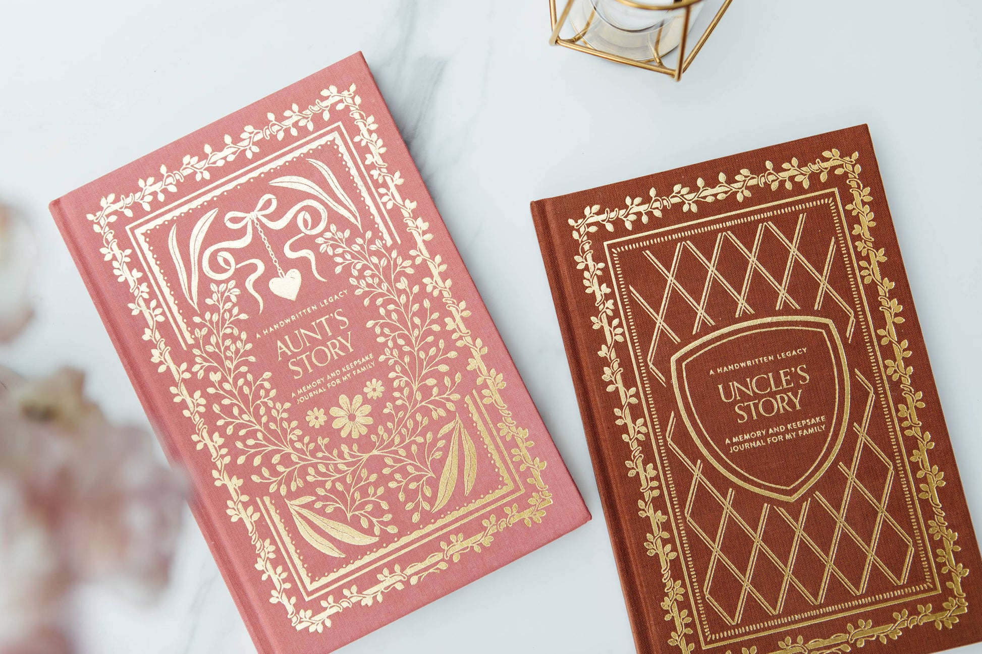Two decorative hardcover books, 'Aunt's Story' in dusty pink and 'Uncle's Story' in dark brown, both featuring ornate gold foil borders and titles, arranged on a light marble surface.