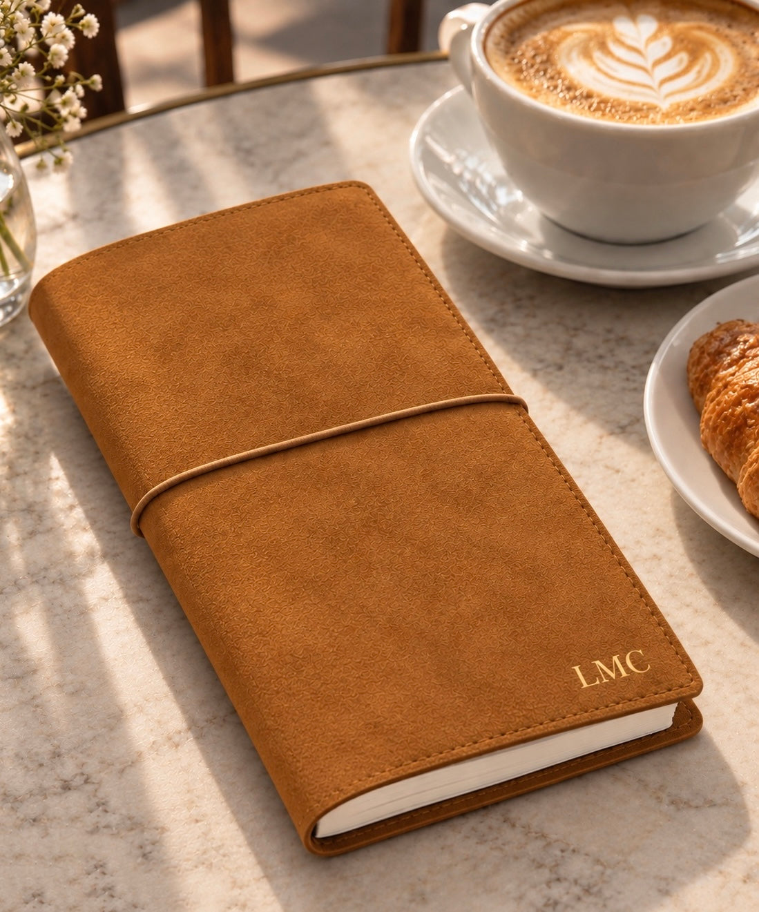 Personalized tan suede journal with elastic closure and gold “LMC” initials, styled beside a latte and croissant on a marble tabletop in warm natural light.