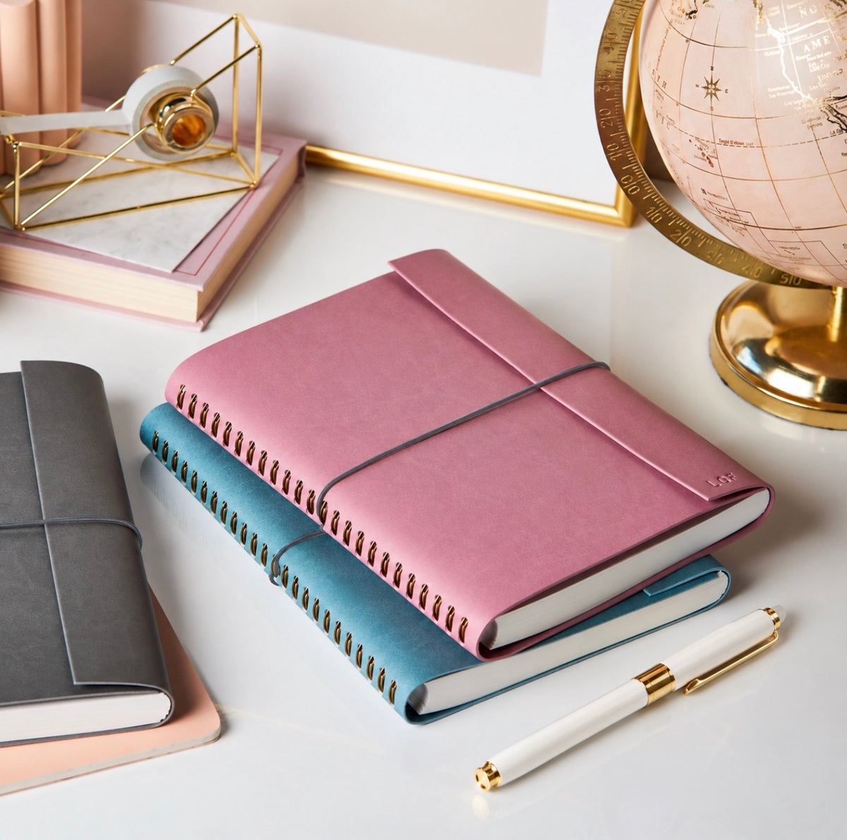 Dusty rose spiral-bound journal stacked on teal and gray journals with a white and gold pen