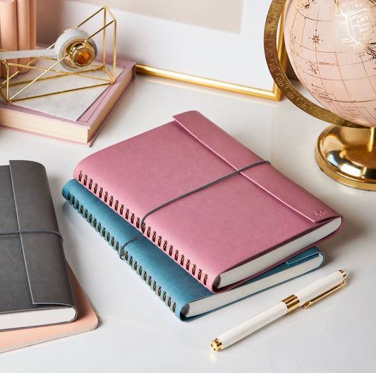 Dusty rose spiral-bound journal stacked on teal and gray journals with a white and gold pen