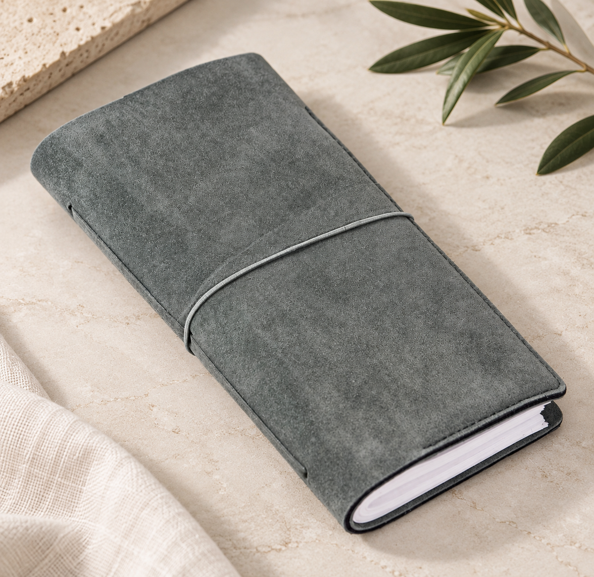 Closed gray-green journal with elastic closure on a neutral stone surface, styled with olive branches and a linen cloth.