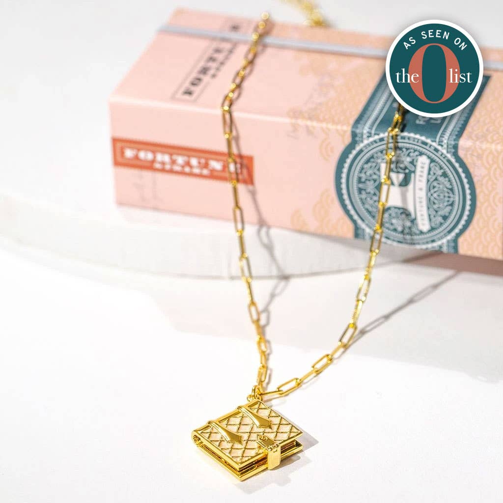 Gold chain necklace with a small vintage-style book pendant displayed on a white surface, with stacked decorative books in the background. A round “As Seen on the O List” badge appears in the upper right.
