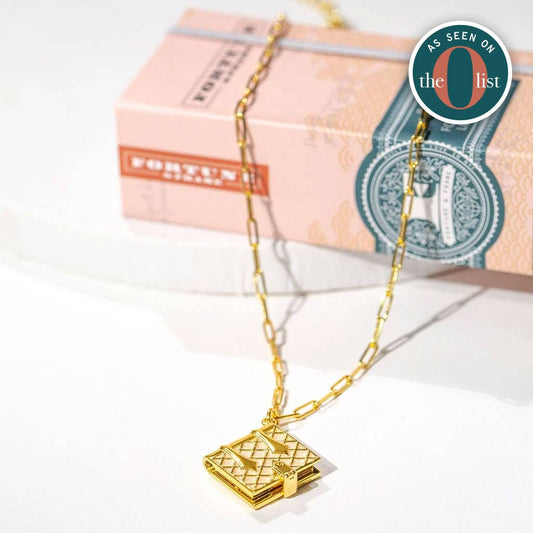 Gold chain necklace with a small vintage-style book pendant displayed on a white surface, with stacked decorative books in the background. A round “As Seen on the O List” badge appears in the upper right.