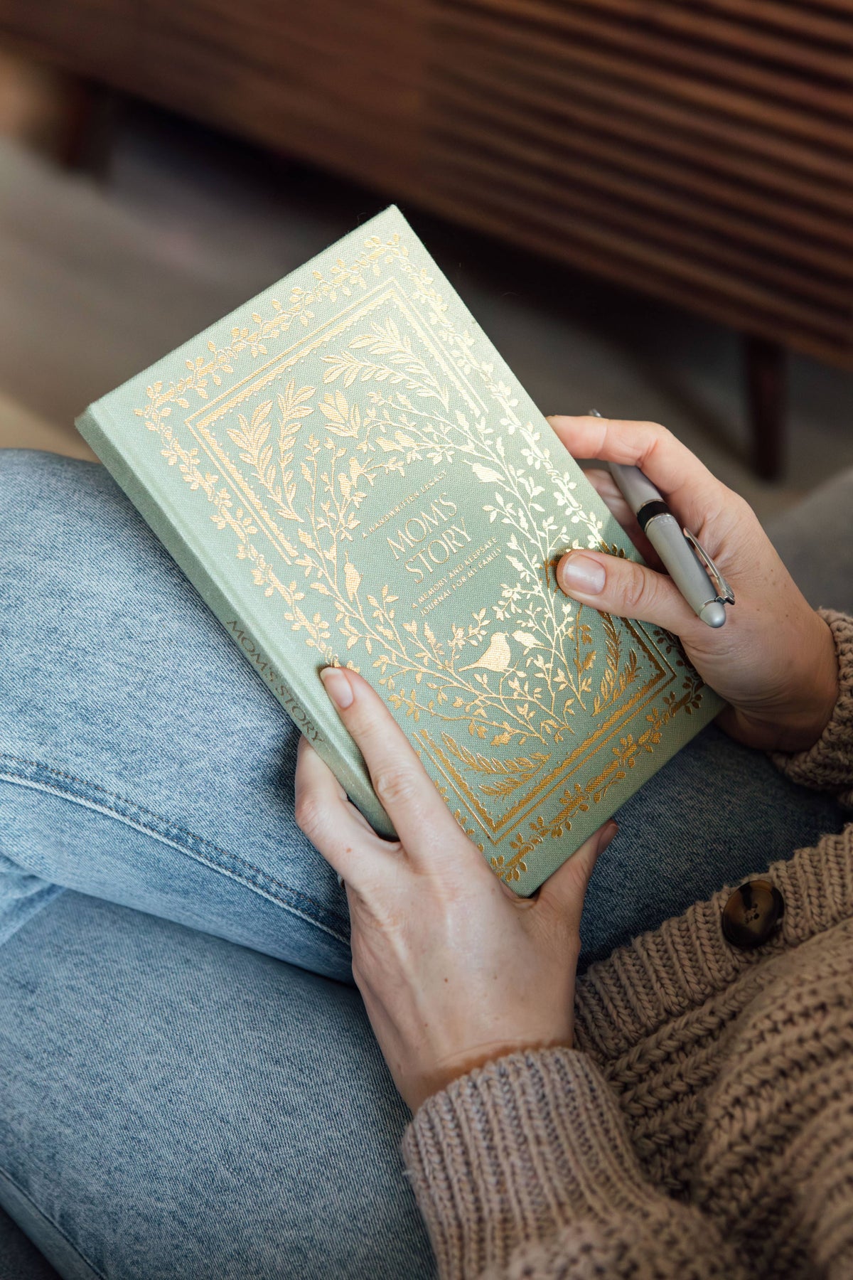 Lifestyle photo of a person holding the green Mom’s Story” keepsake journal with gold botanical detailing and a pen.