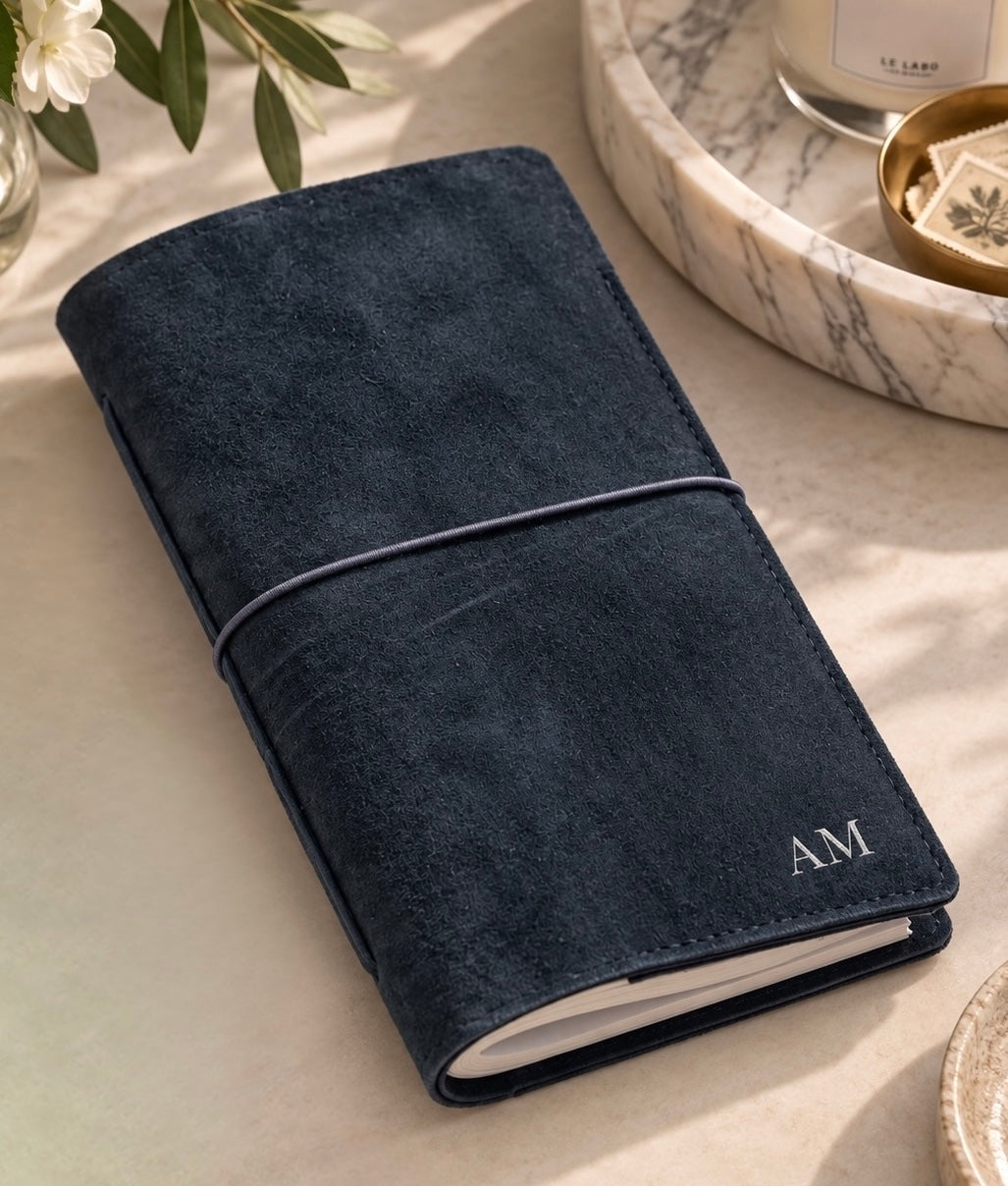 Personalized navy journal with elastic closure and white “AM” initials, styled on a warm neutral tabletop with soft natural light, greenery, a marble tray, and a candle.