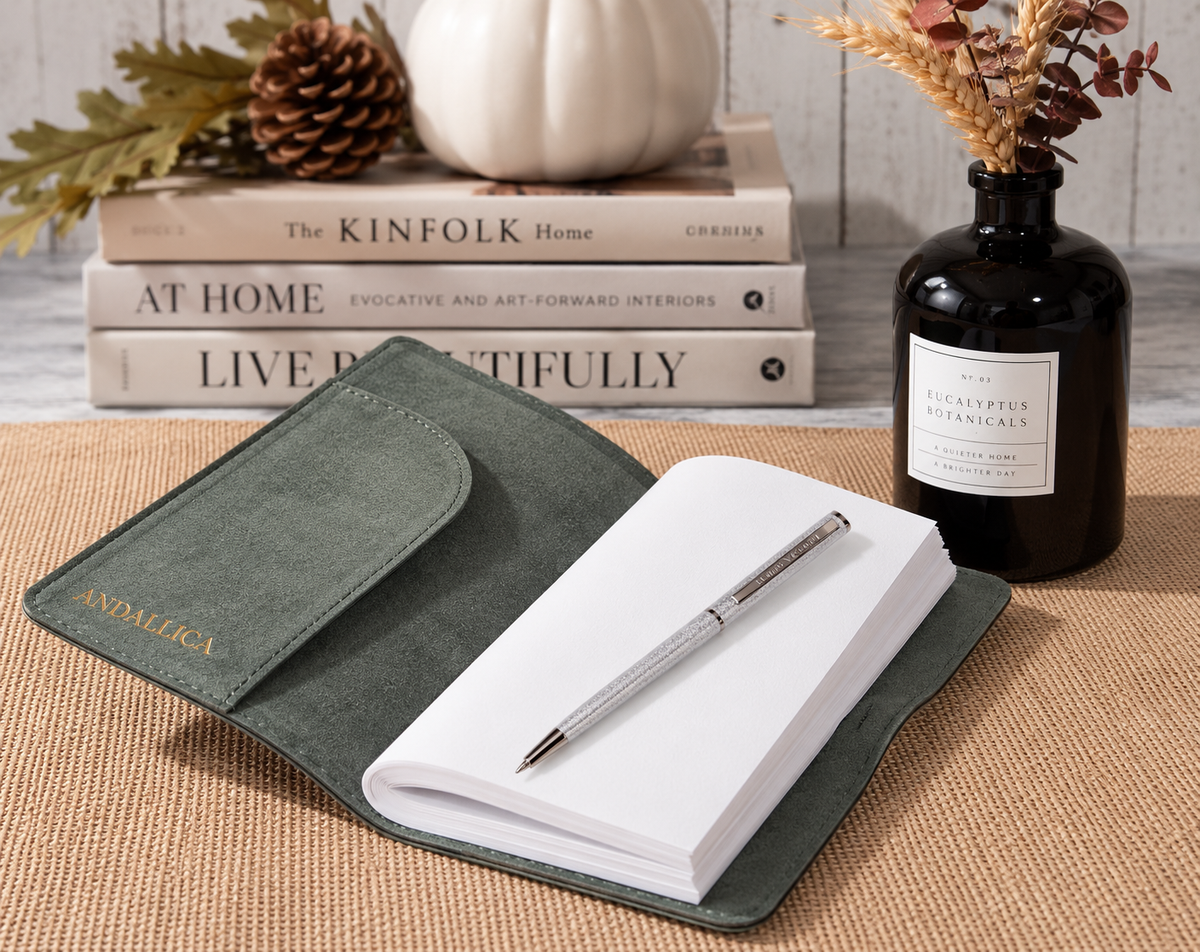 Open sage green leather journal with a silver pen, styled on a woven tabletop beside stacked books and a botanical vase.