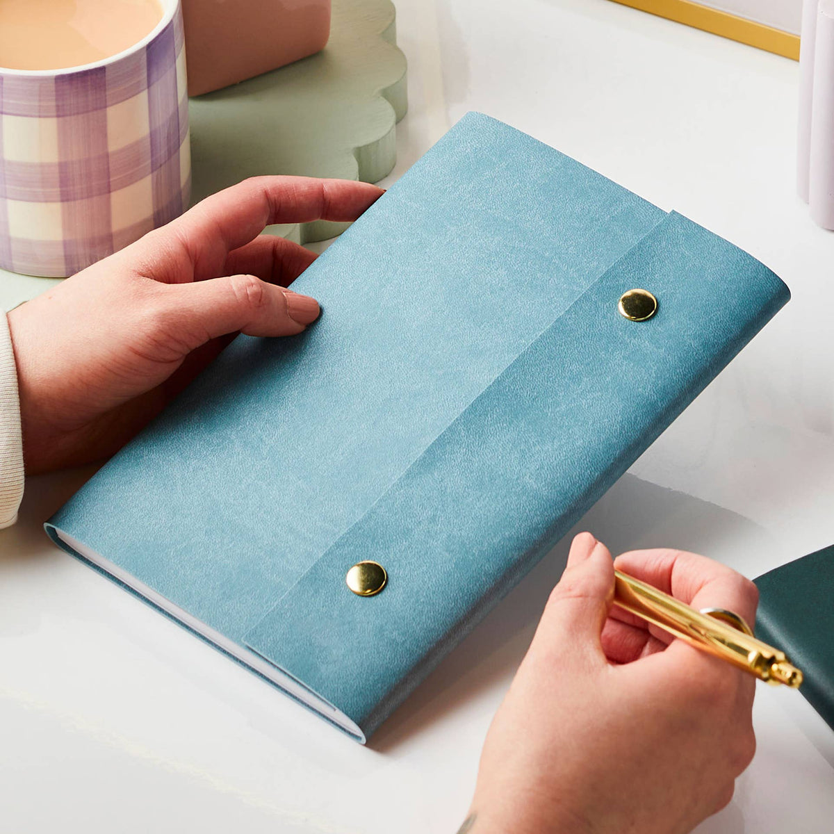 Teal leather journal with gold pen held on a white desk