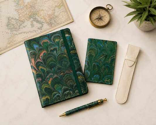 A vintage-inspired travel journal with a rich teal and green peacock-feather pattern, paired with a matching smaller notebook, a coordinating pen, and a cream pen sleeve. A vintage map, brass compass, and potted greenery create an elegant travel-themed still life.